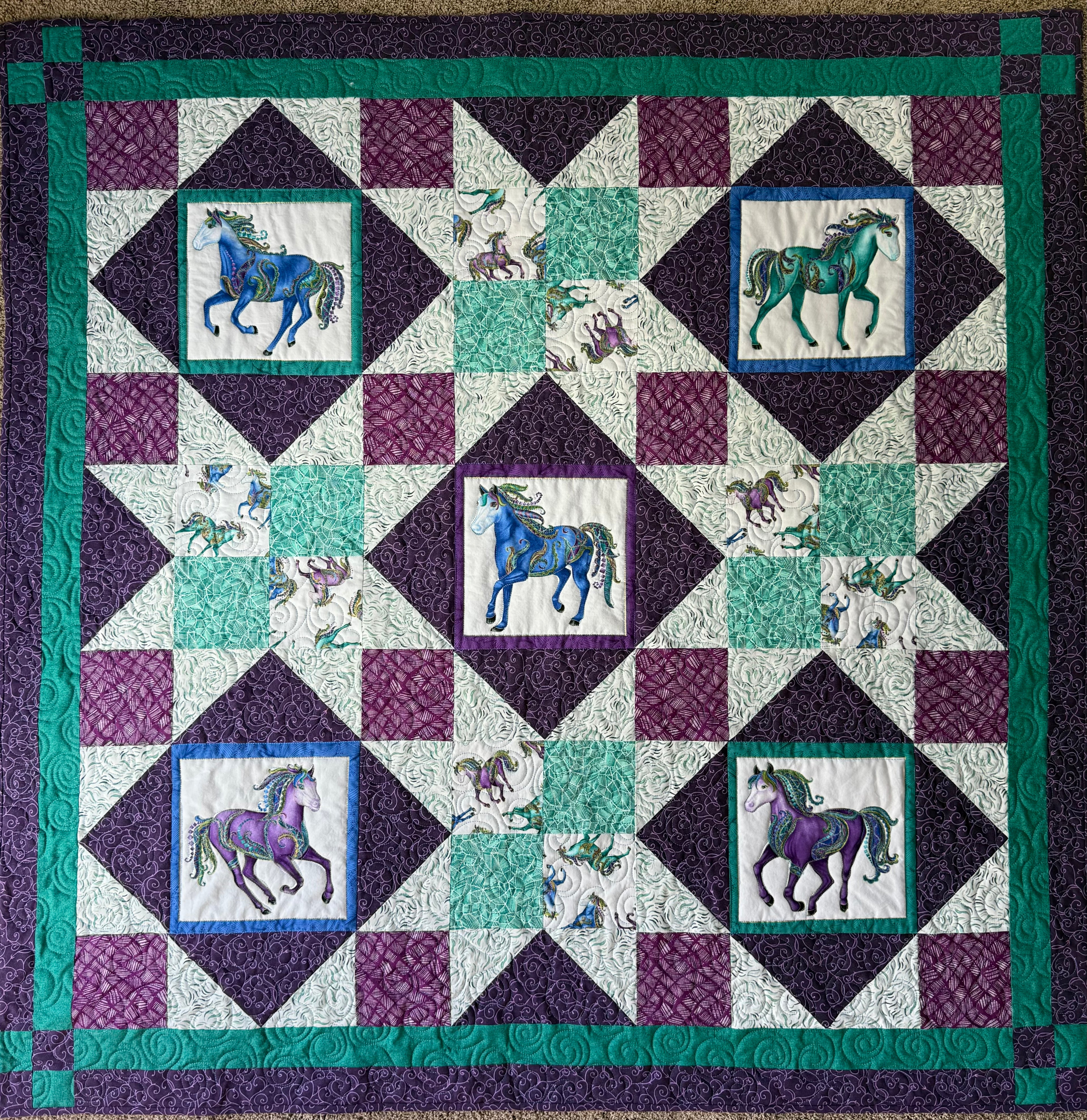 Handmade quilt featuring a repeating pattern of horses in shades of blue and purple. The quilt has a patchwork design with squares and diamonds in teal, white, and purple. The horses are detailed with decorative elements. 