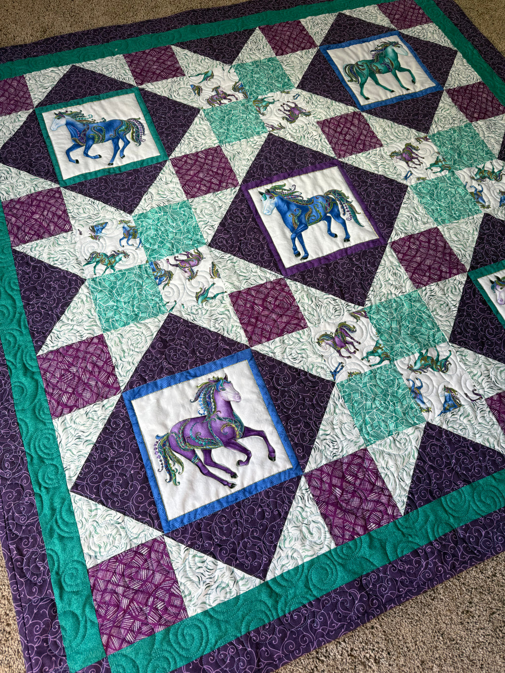 A handmade quilt featuring a repeating pattern of horse illustrations in squares. The quilt has a purple, teal, and white color scheme. The horses are in various colors, including blue, purple, and green. The quilt is bordered with teal fabric.
