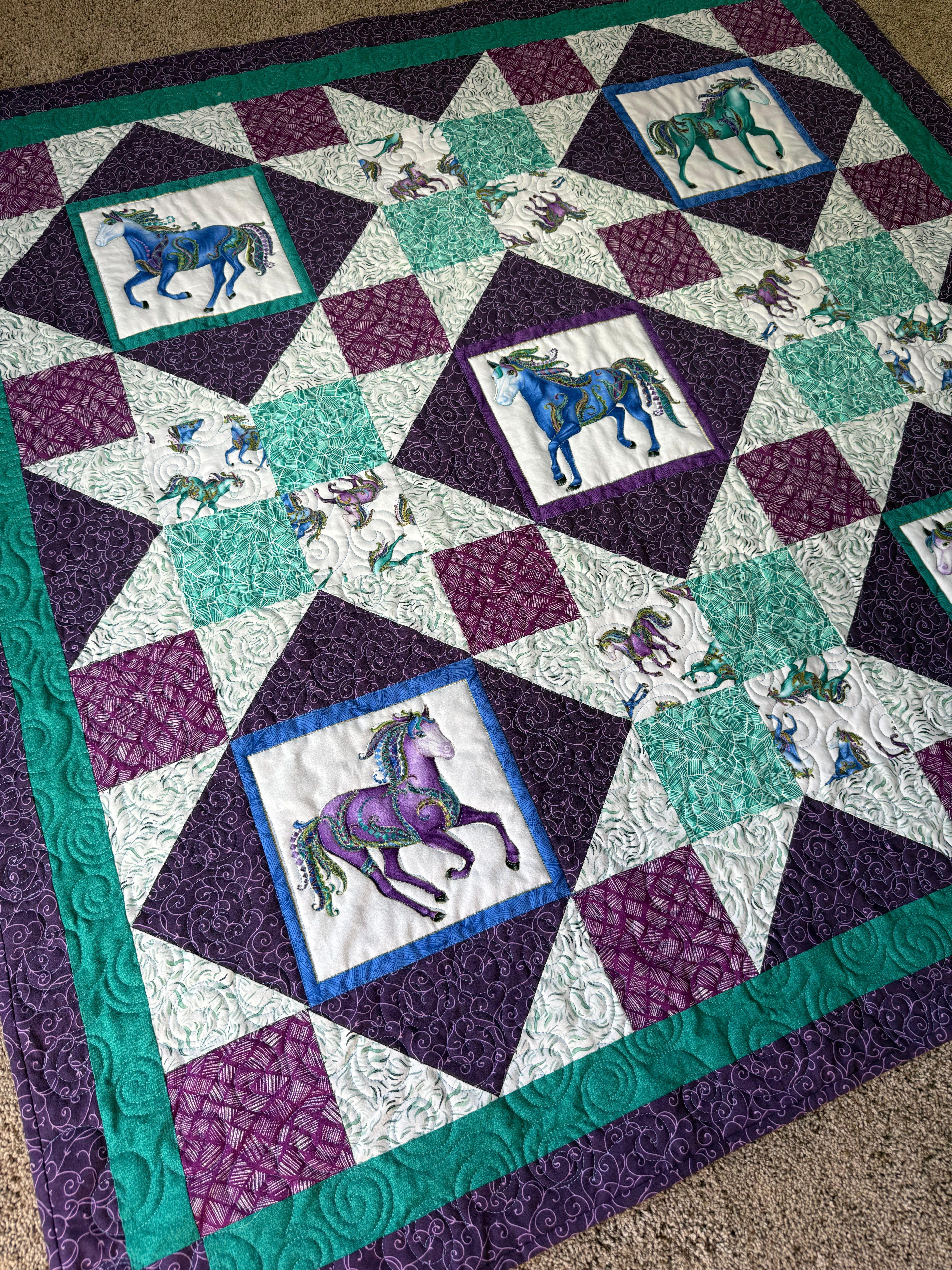 A handmade quilt featuring a repeating pattern of horse illustrations in squares. The quilt has a purple, teal, and white color scheme. The horses are in various colors, including blue, purple, and green. The quilt is bordered with teal fabric.