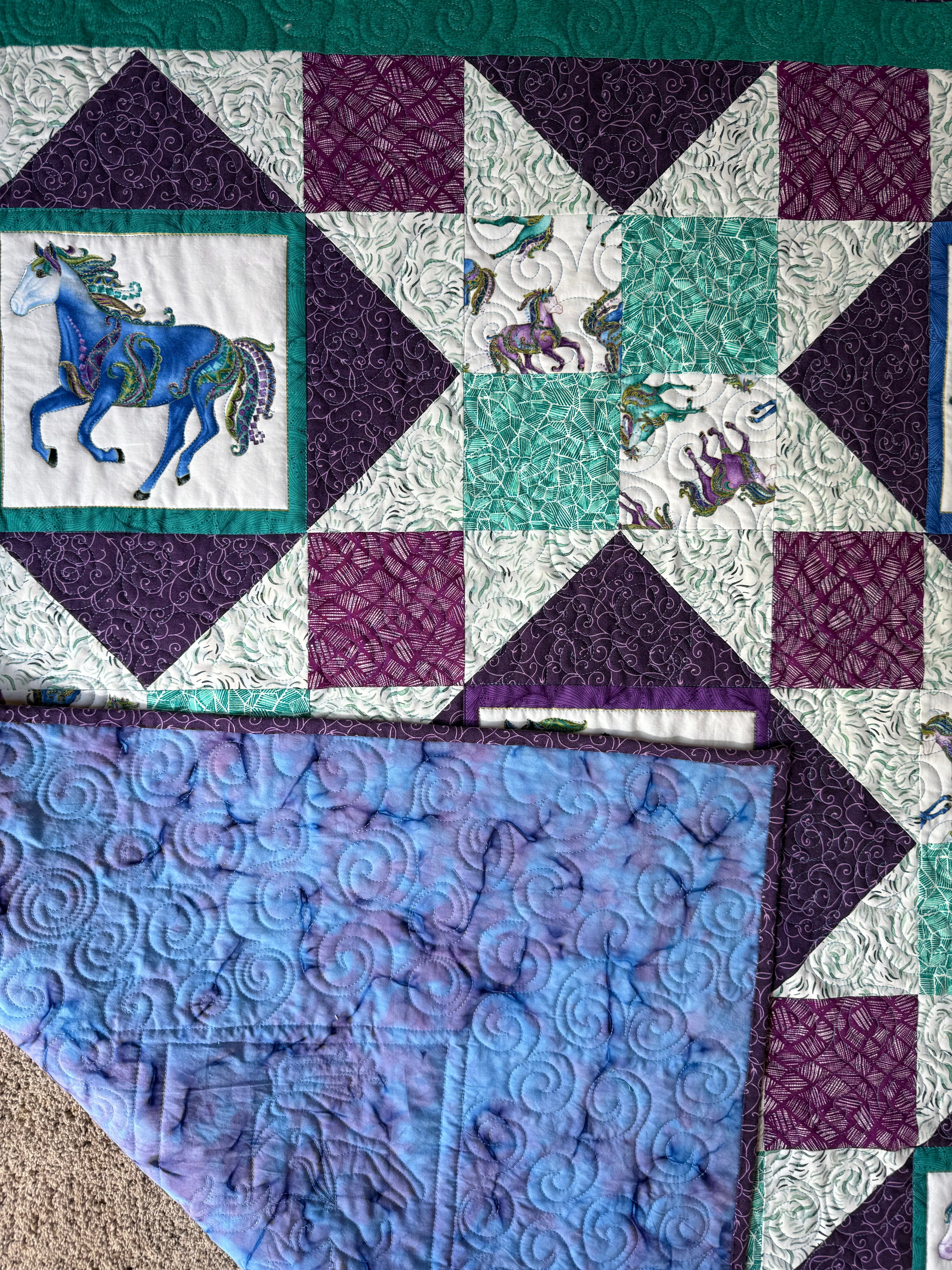 A handmade quilt featuring a horse design. The quilt showcases a patchwork design with squares and triangles in shades of purple, teal, and white. The backing is a blue and purple tie-dye pattern. The quilt is made of cotton.