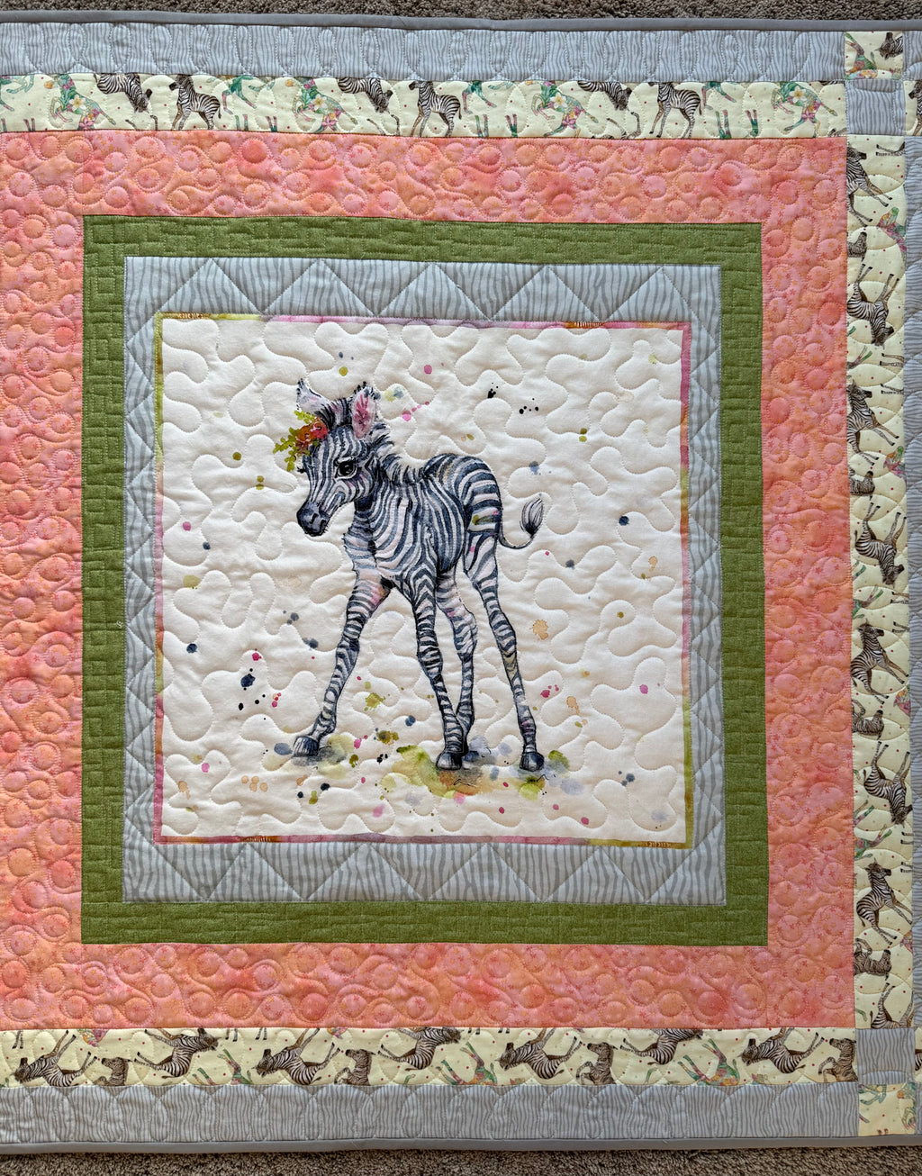 A handmade quilted baby blanket featuring a central image of a zebra foal with a floral crown. The quilt has a square design with a white background, surrounded by layers of pink, green, and gray patterned borders. The outer border has a zebra print. 
