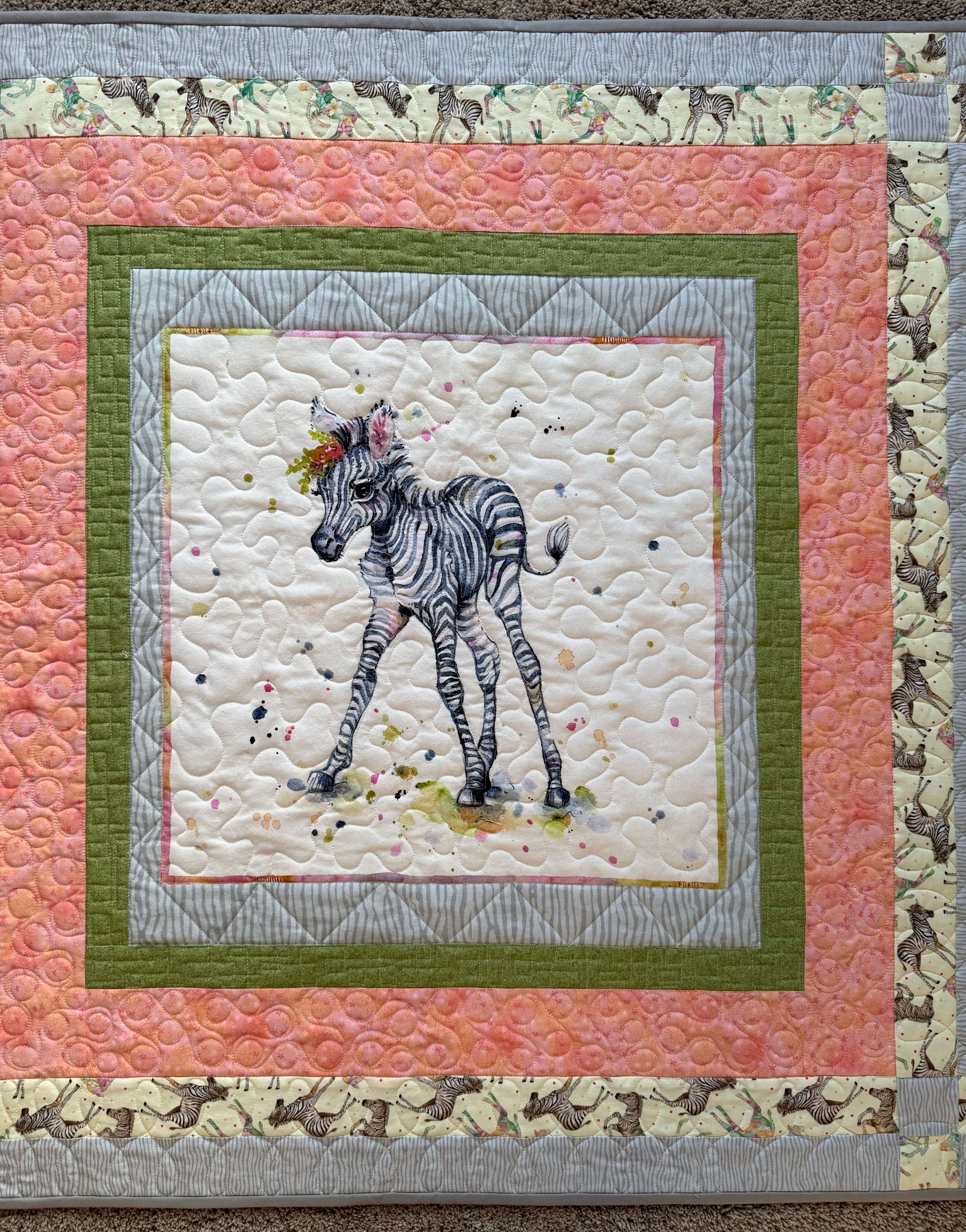 A handmade quilted baby blanket featuring a central image of a zebra foal with a floral crown. The quilt has a square design with a white background, surrounded by layers of pink, green, and gray patterned borders. The outer border has a zebra print. 
